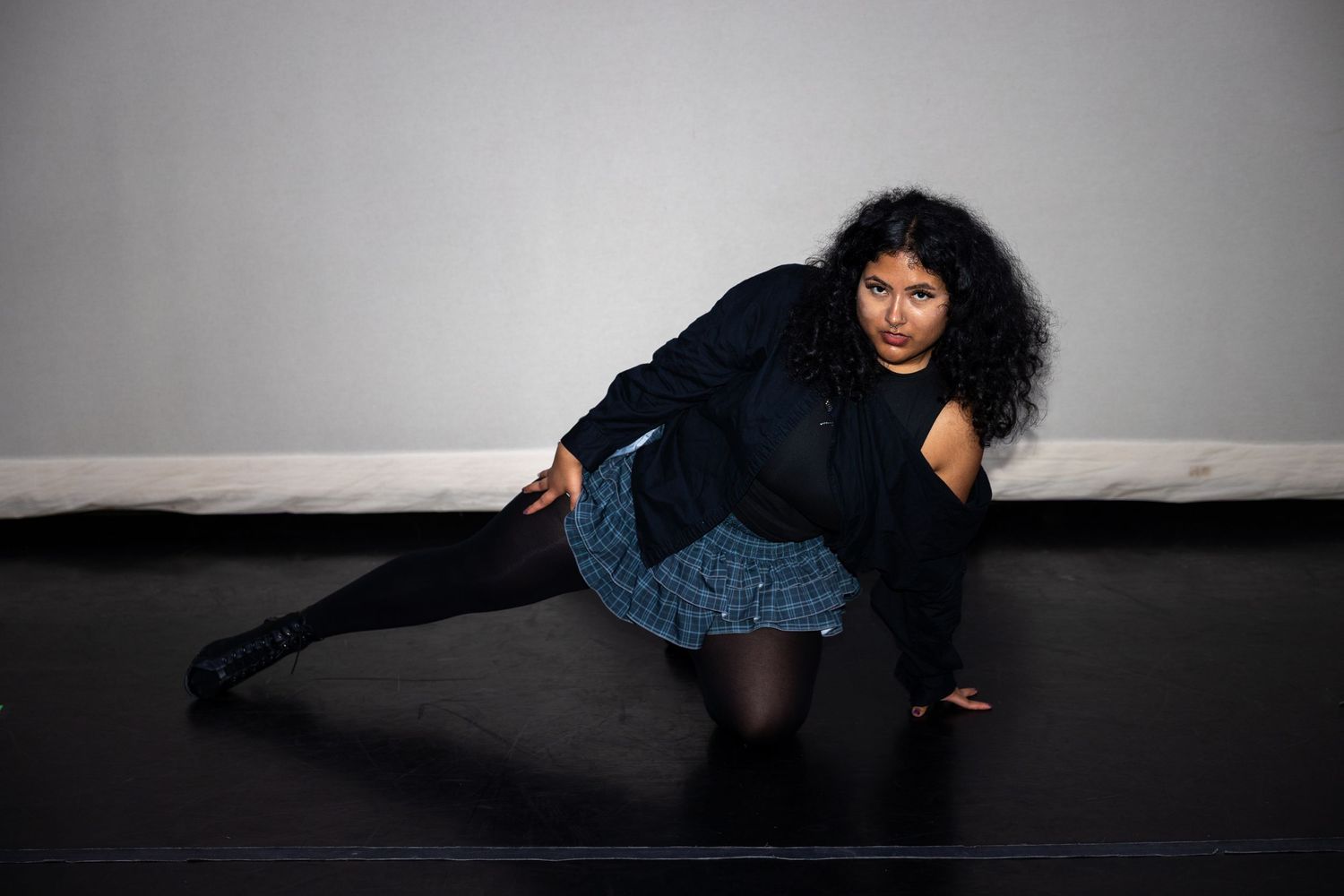 A dancer in a blue plaid skirt and black jacket poses low on a dark studio floor.