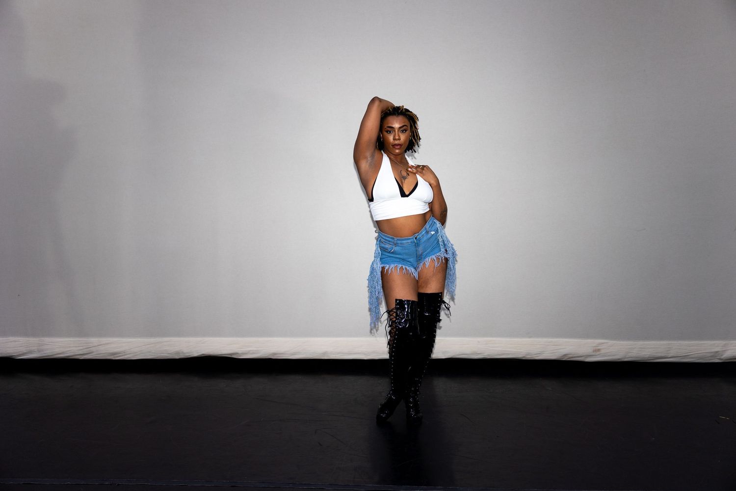 Dancer in white sports bra and denim skirt strikes confident pose wearing thigh-high boots on stage.