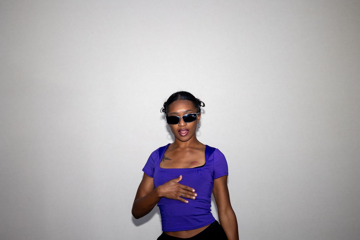 Stylish young Black woman wearing purple crop top and sunglasses posing confidently against a white background.