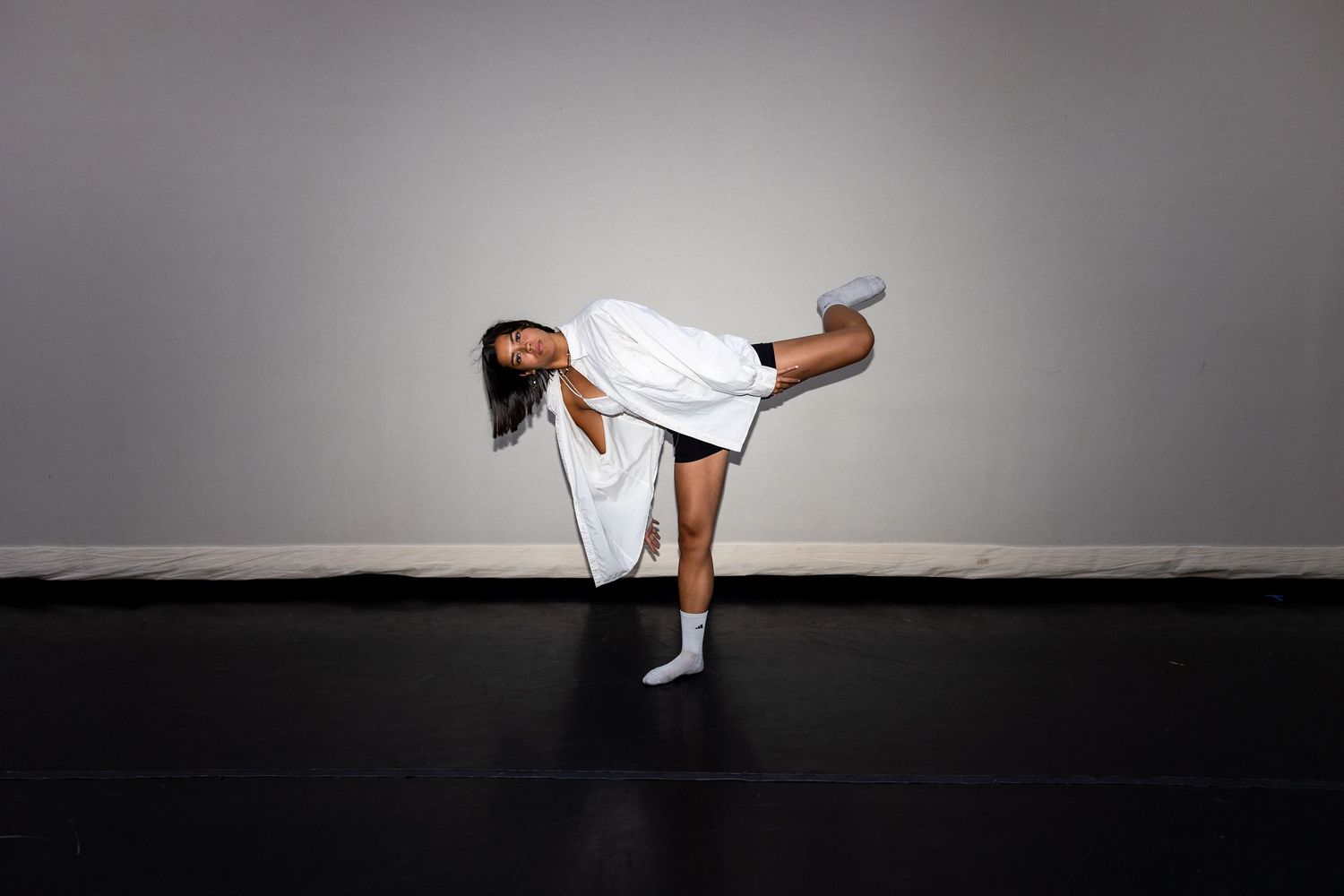 A dancer in a white outfit performs a dramatic side kick lean on a dark studio floor.