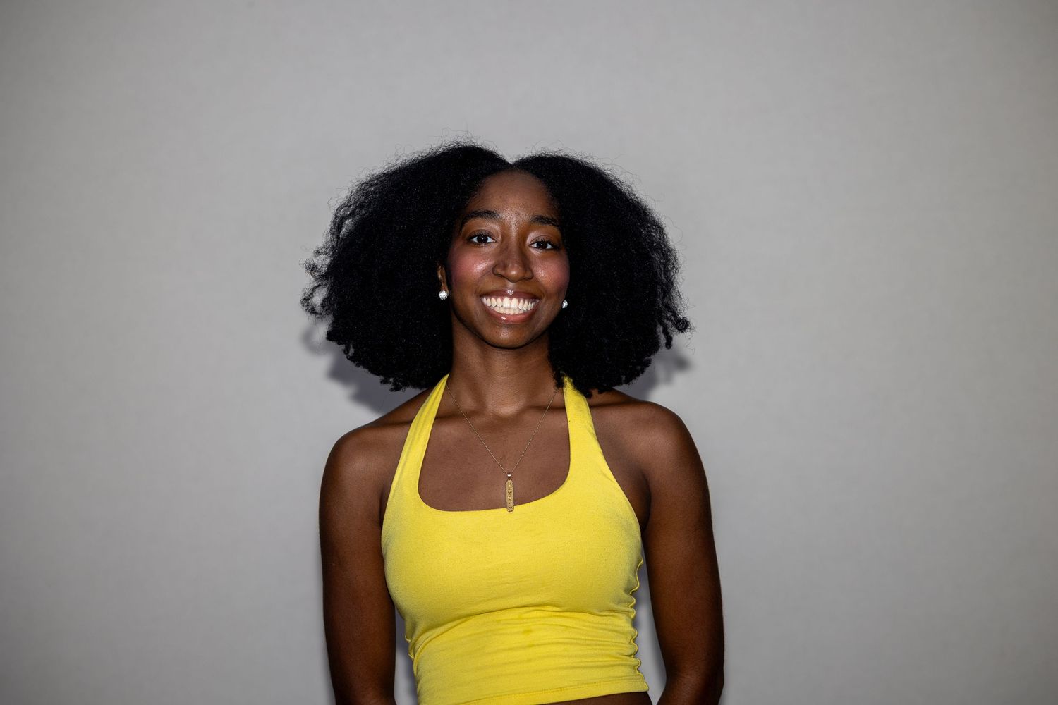 Smiling woman with natural afro hair wearing a yellow halter top against a neutral background.