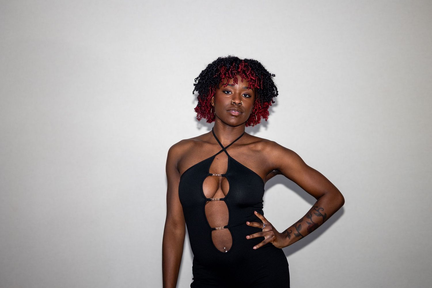 Black woman with red curly hair in a black cutout halter bodysuit poses with hand on hip.