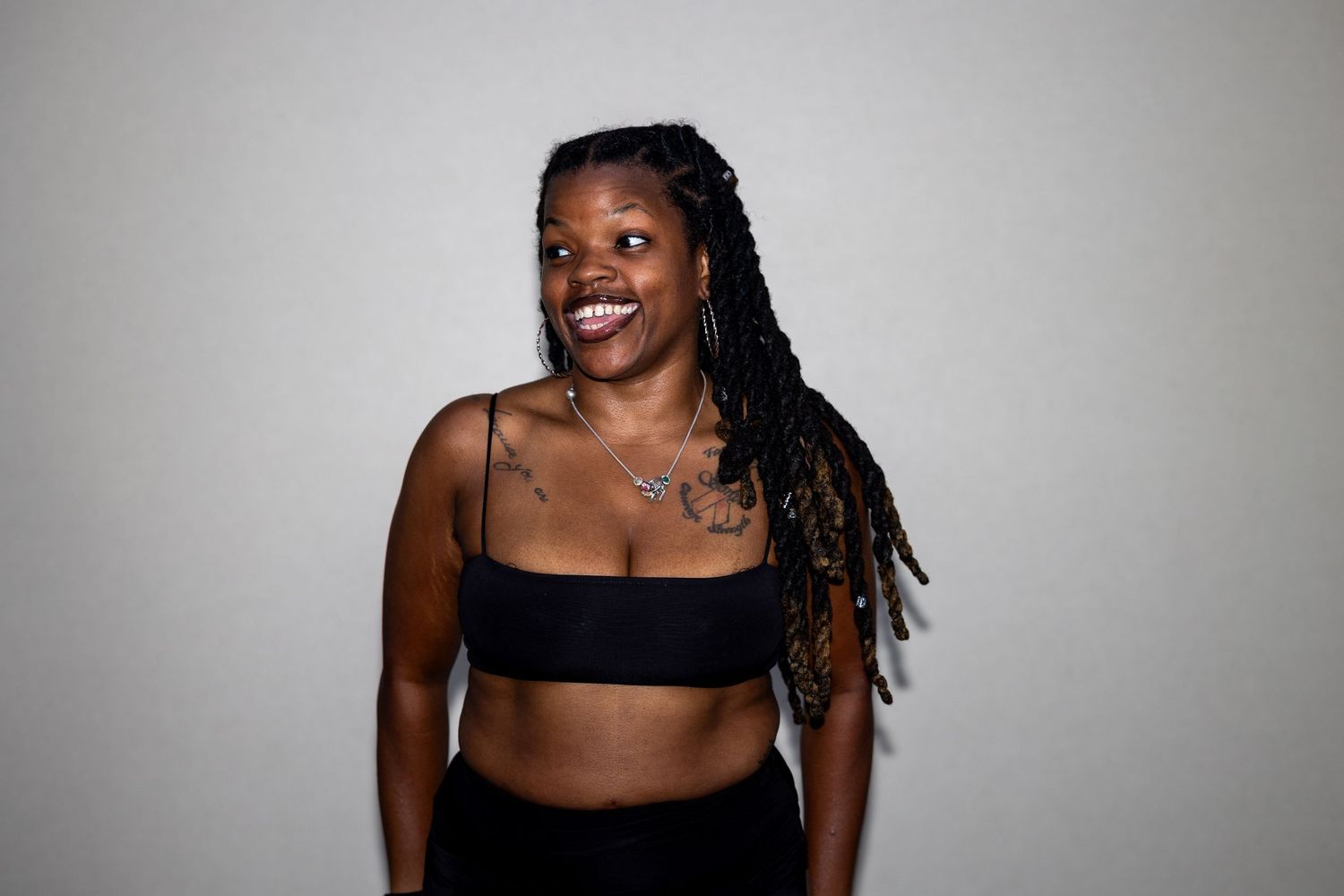 Smiling woman with long braids wearing black crop top and pants with visible tattoos against white background.