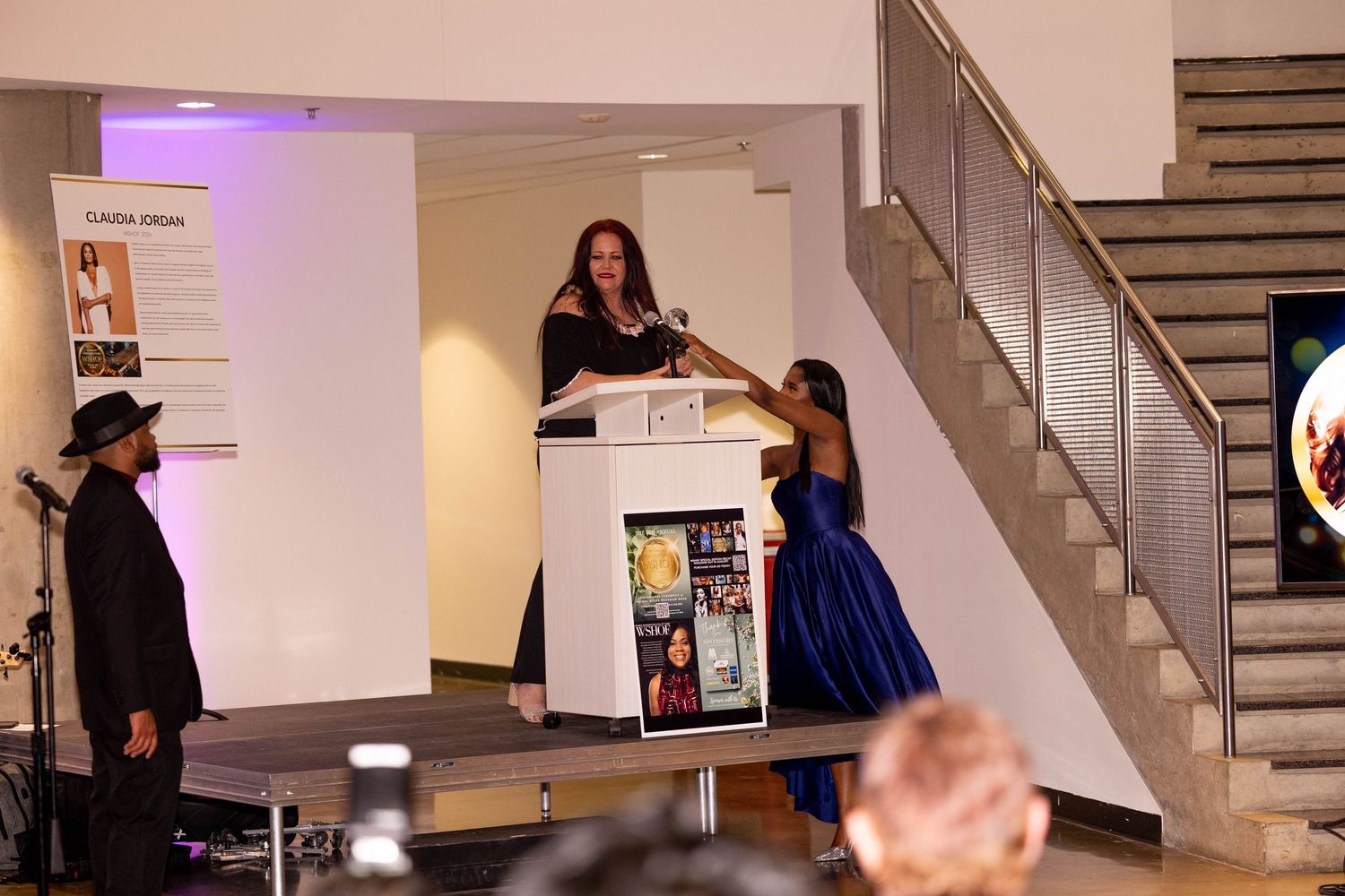 Woman speaking at podium on stage during formal event, with guests in elegant attire and staircase in background.
