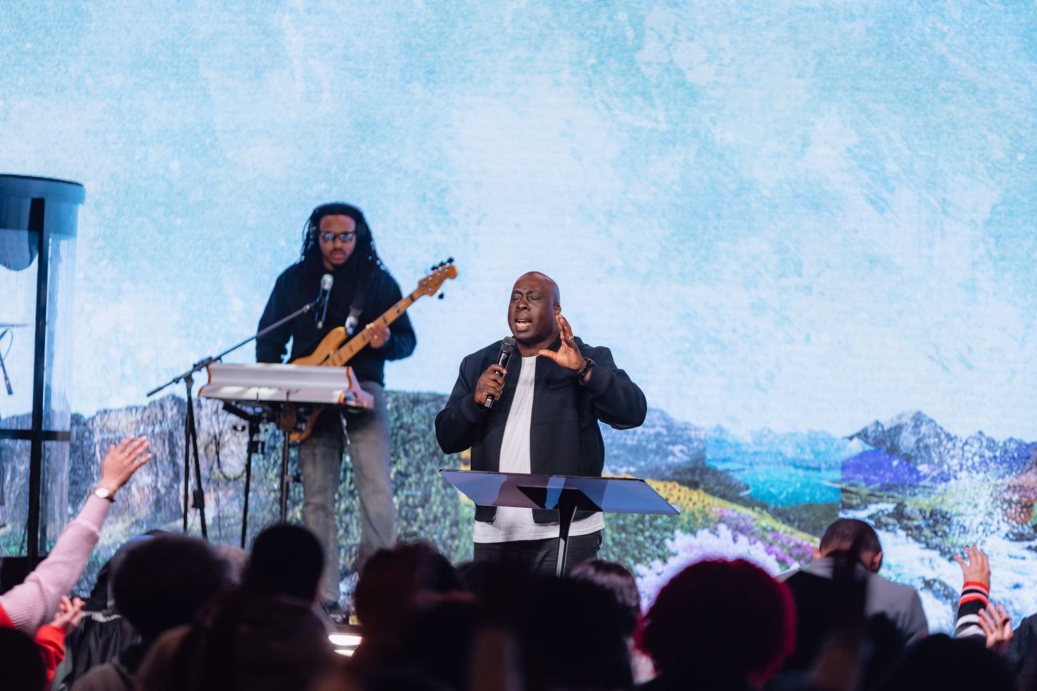 Musicians perform on stage at an outdoor festival with mountains visible in the background.
