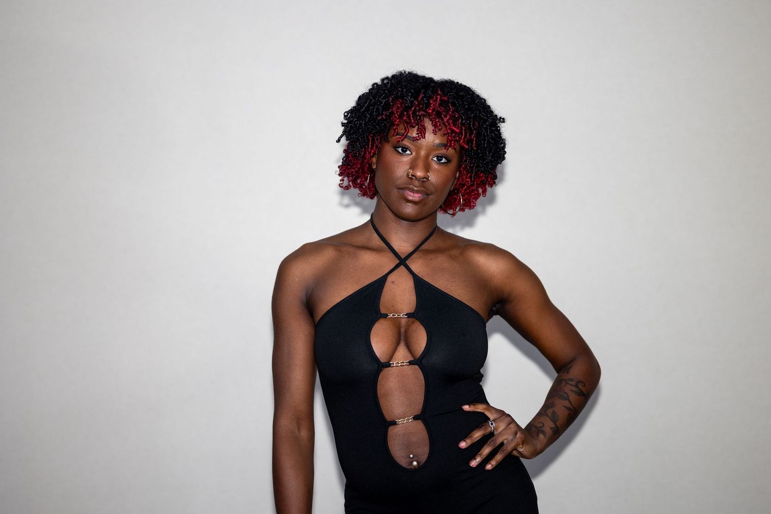 Woman in black cutout halter dress with red-tipped curly hair posing with hand on hip against white background.
