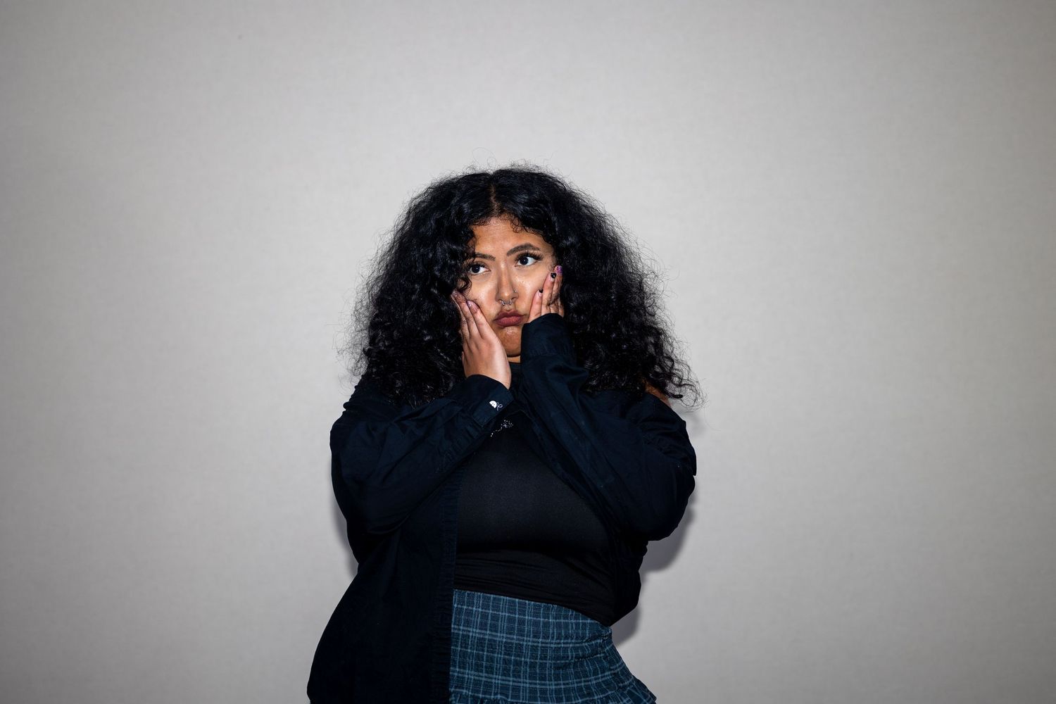 Young woman with curly hair in a black top and plaid skirt, posing with hands on face against a gray background.