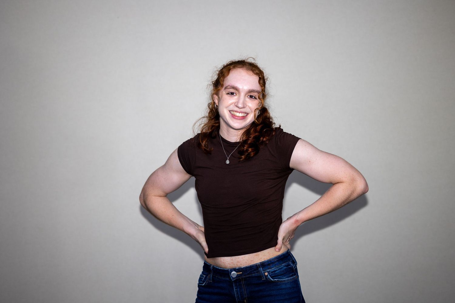 Young woman with auburn hair in a black crop top and jeans smiles with hands on hips.