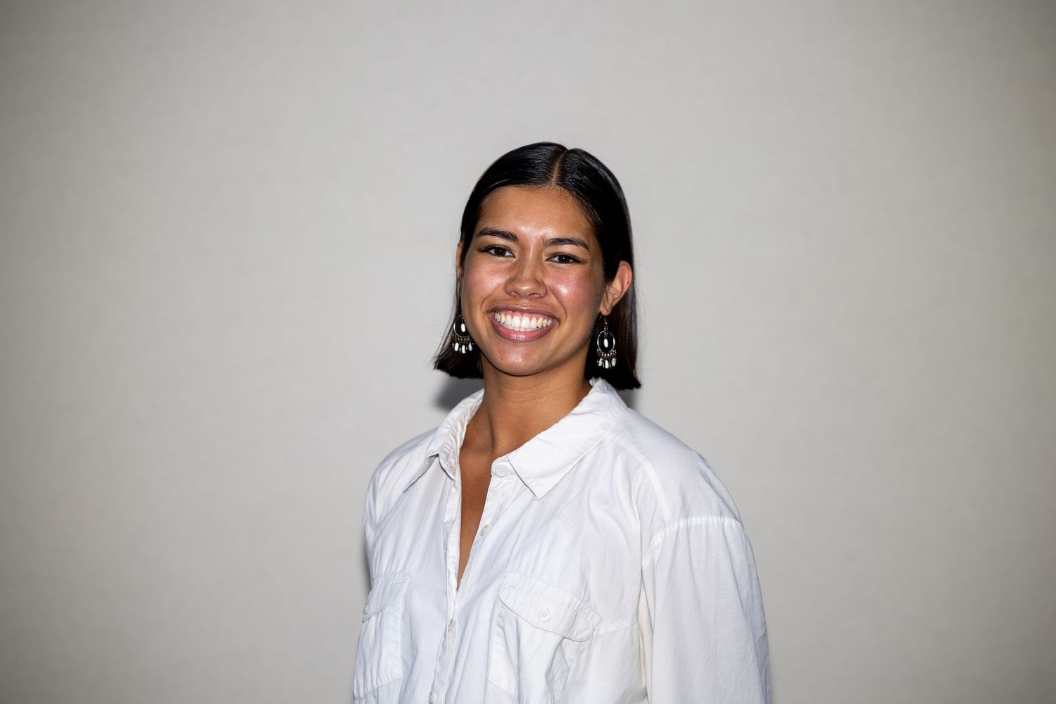 Smiling woman in white button-up shirt with black earrings poses against a neutral background.