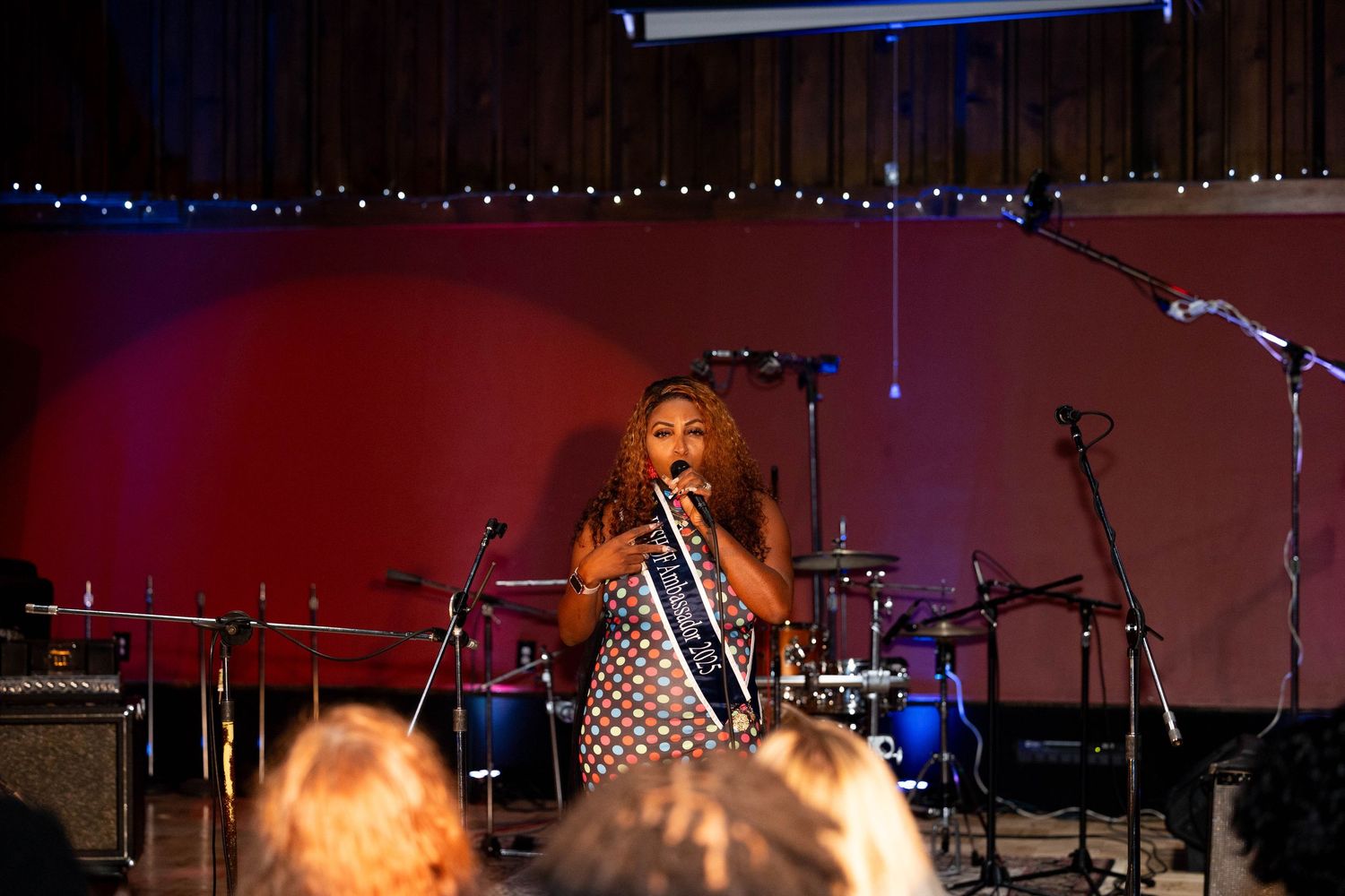 A female singer performs on stage with a microphone, backed by drums and instruments in a warmly lit venue.