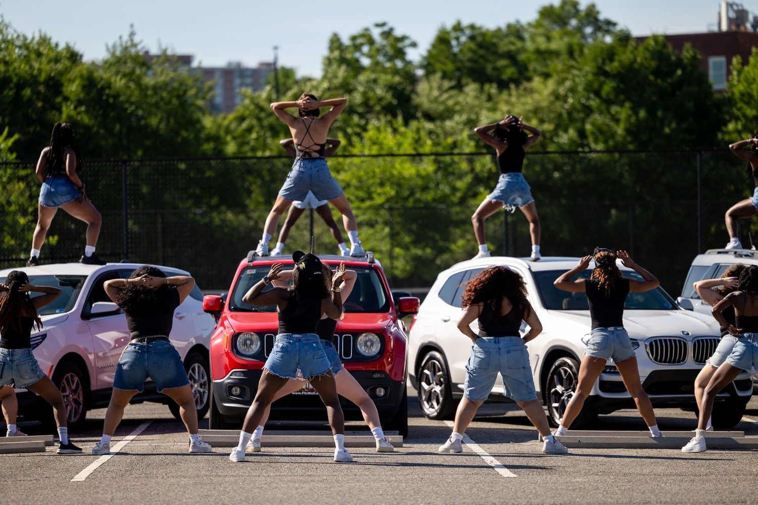 A dance team performs a synchronized routine in a parking lot, with one dancer lifted high above the group.