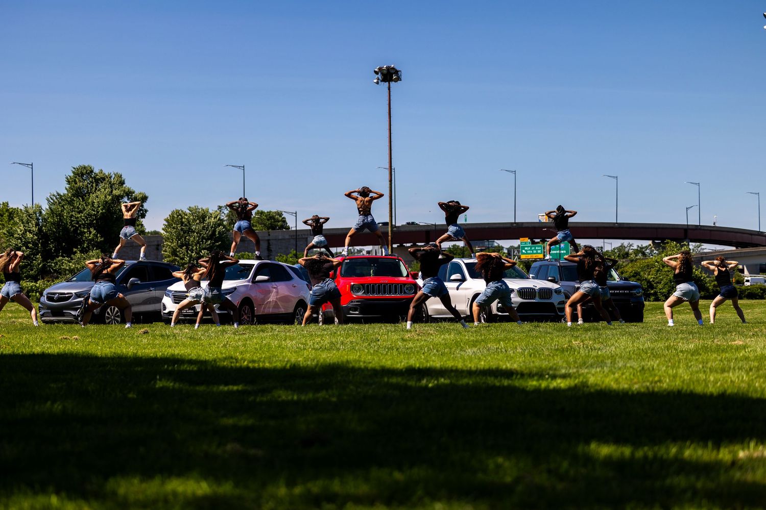 Dancers perform synchronized acrobatic moves on a grassy field with colorful cars and trees in the background.