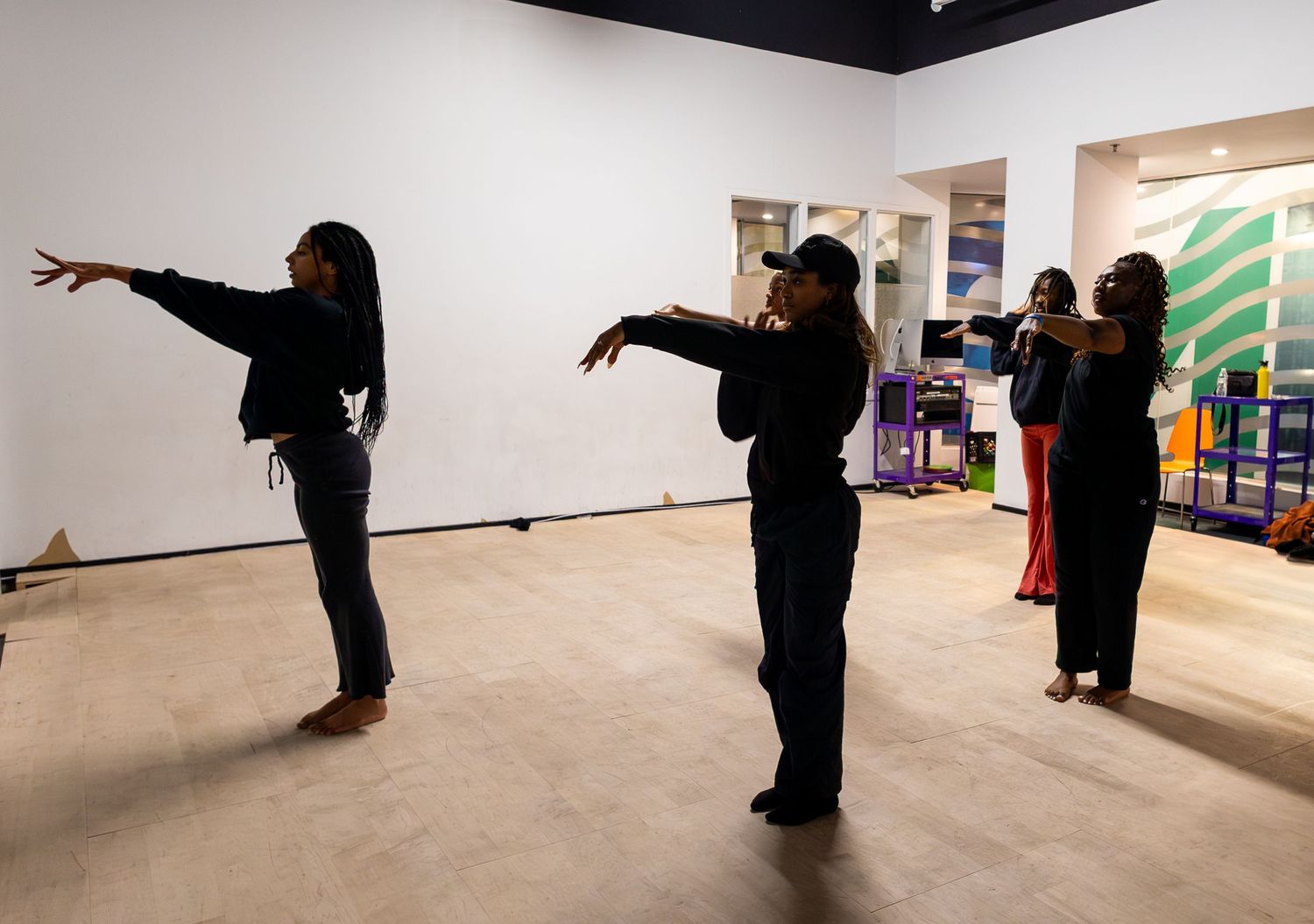 Four women practice dance moves in a bright, open gallery space with white walls and colorful artwork.