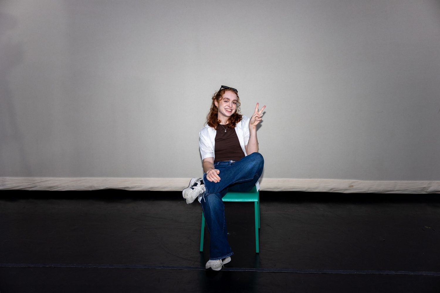 A smiling young woman in jeans sits on a green chair giving a peace sign in a dance studio.
