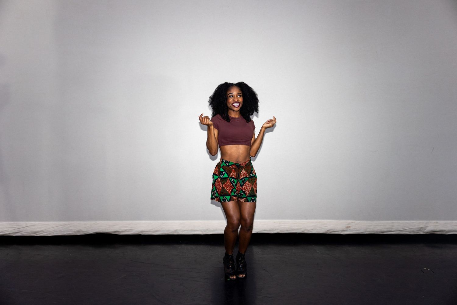 Joyful Black woman in African print skirt and maroon crop top laughing with hands raised on stage.
