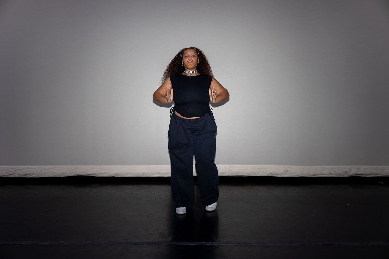 A woman in a black tank top and wide-leg jeans poses confidently against a gray background.