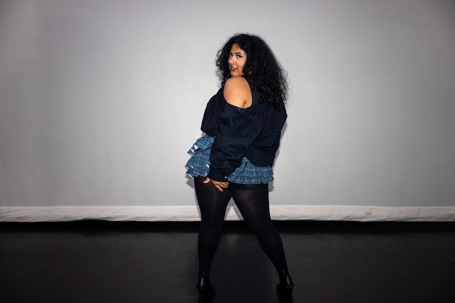 Curvy woman in denim ruffle skirt and navy jacket poses confidently against gray studio backdrop.