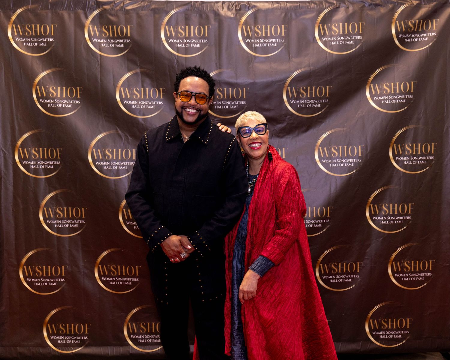 Two elegantly dressed individuals posing together at a WSHOF event backdrop, woman in red outfit with decorative mask.