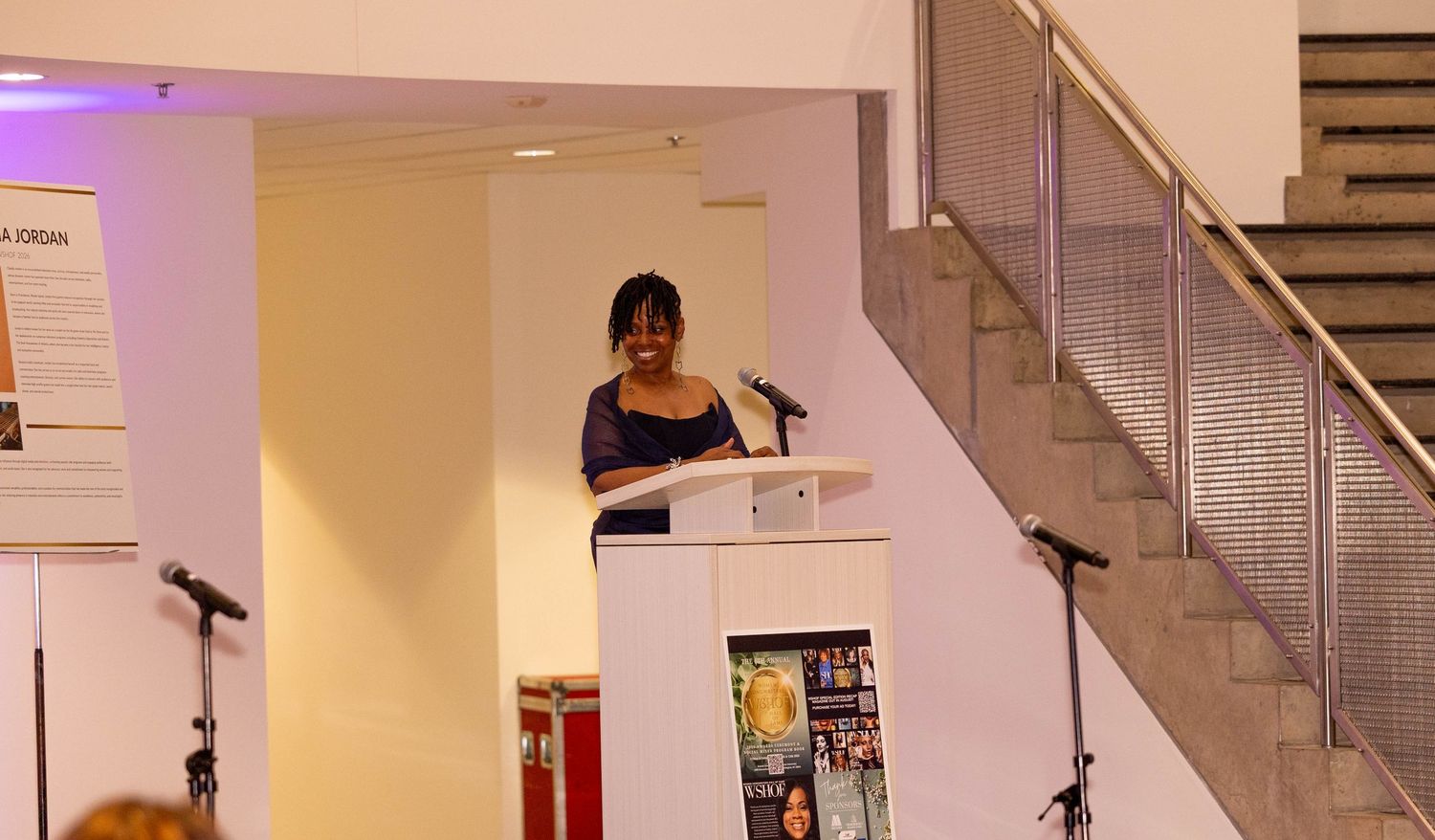 Woman presenting at a white podium in a modern venue with a staircase and display boards visible.