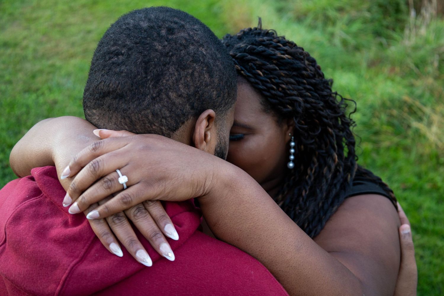 A joyful woman with braided hair hugs her partner, showcasing a sparkling engagement ring in a park setting.