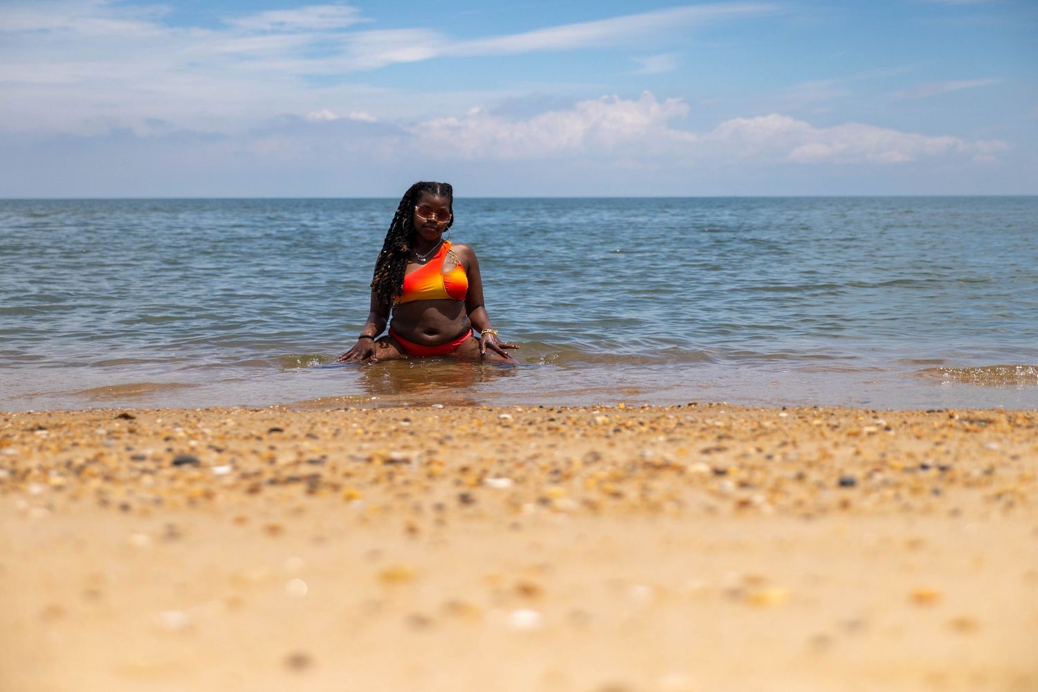 Woman in orange bikini sitting on sandy beach shore with calm ocean waves and blue sky behind her.