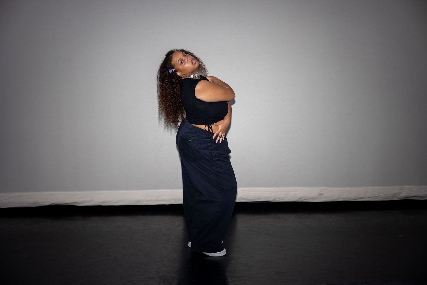 Young woman in black crop top and wide-leg jeans posing confidently against a gray studio wall.