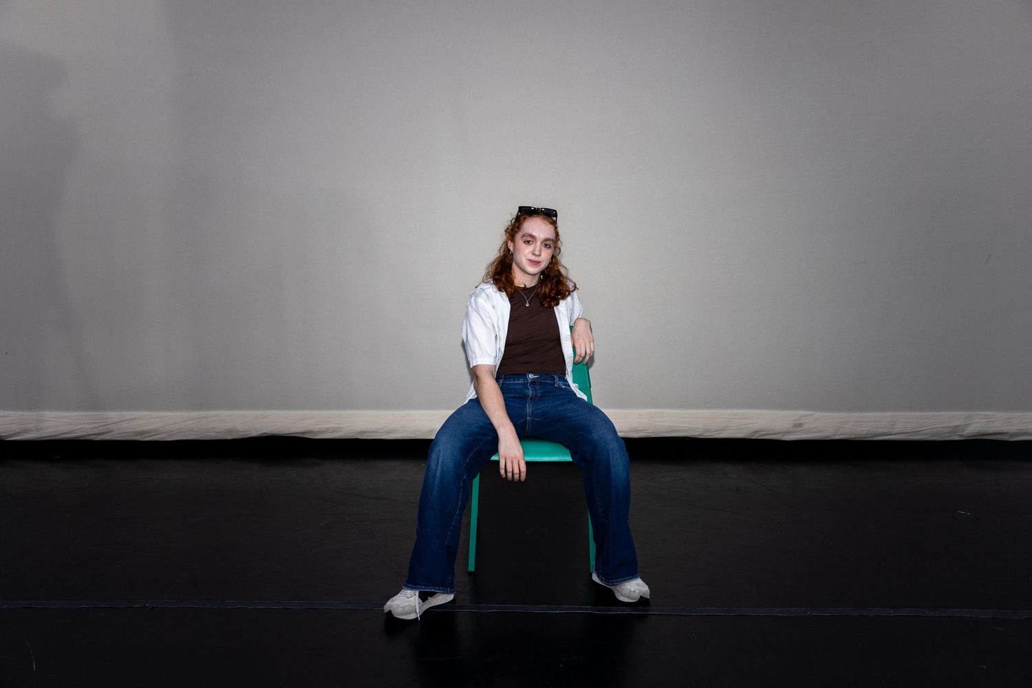 A young woman in jeans and a white shirt sits casually on a black box against a gray background.