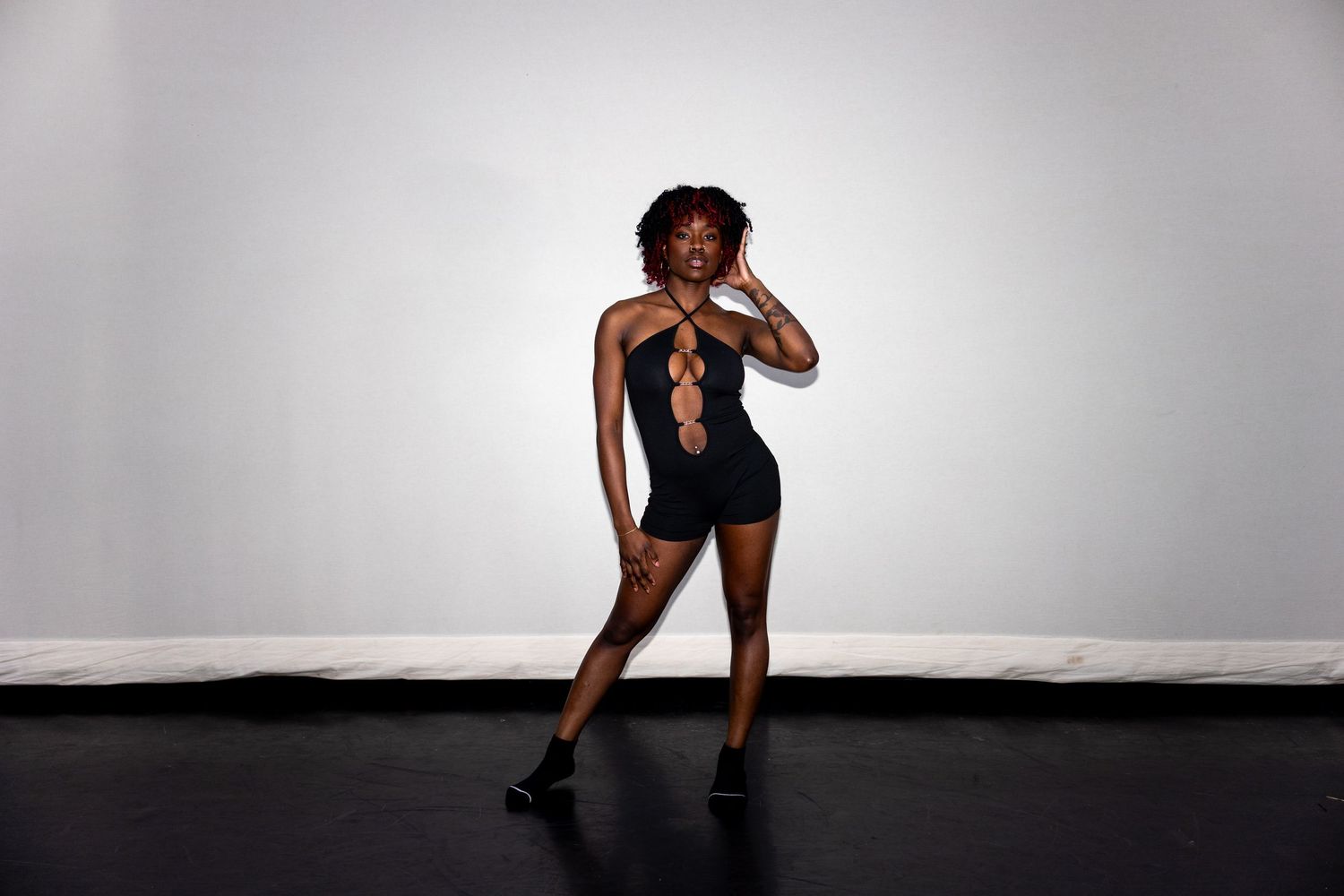 A dancer in a black cutout bodysuit strikes a pose on a dark studio floor.