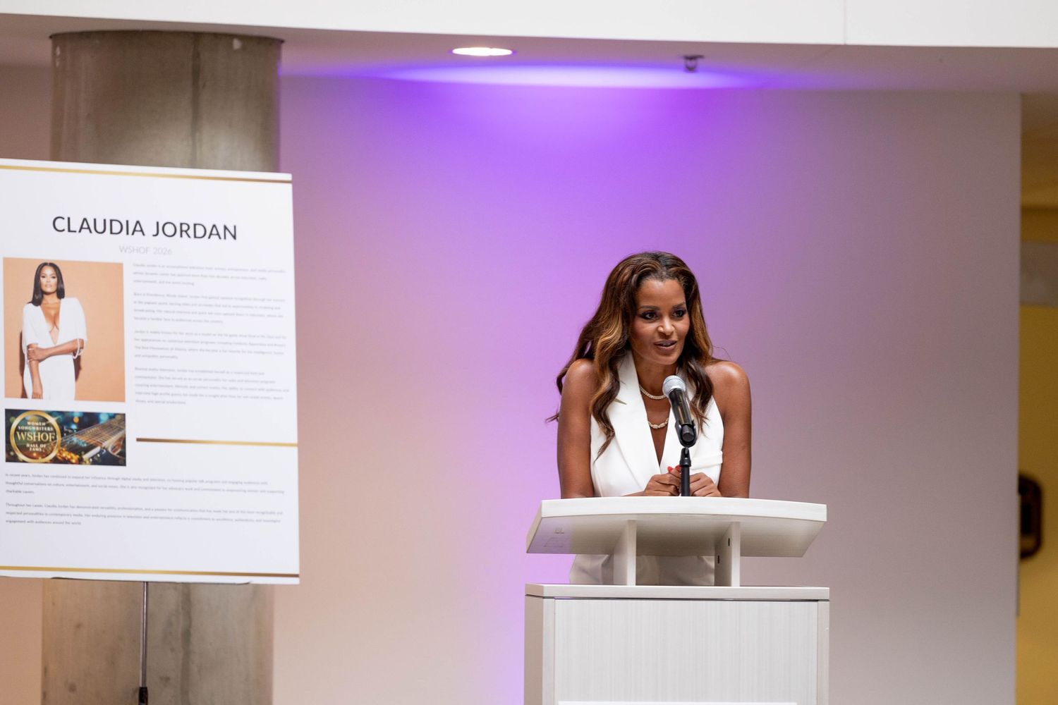 Claudia Jordan speaking at podium during formal event with biography display board visible behind her.