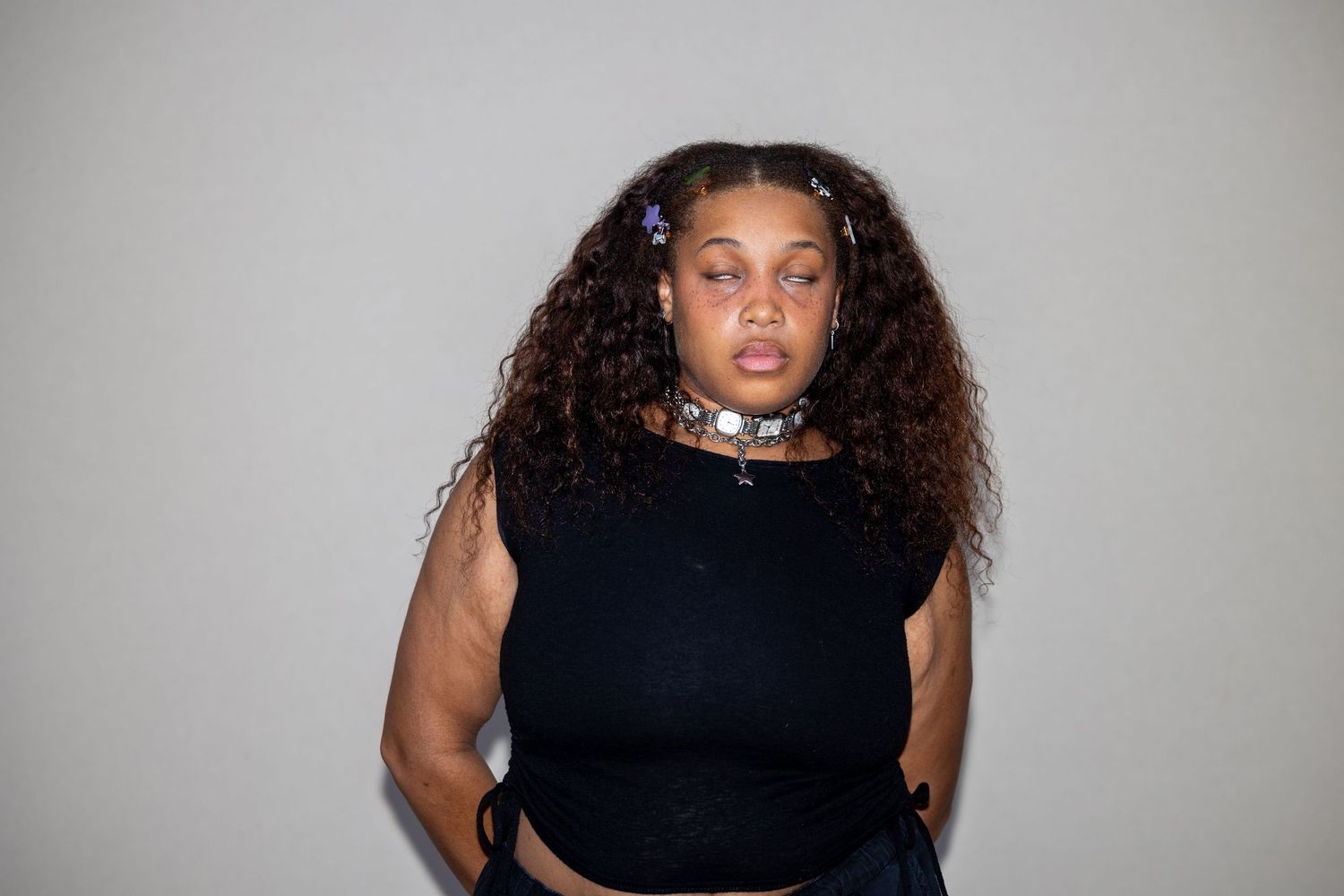 Woman with curly hair wearing black sleeveless top and choker necklace stands against gray wall.