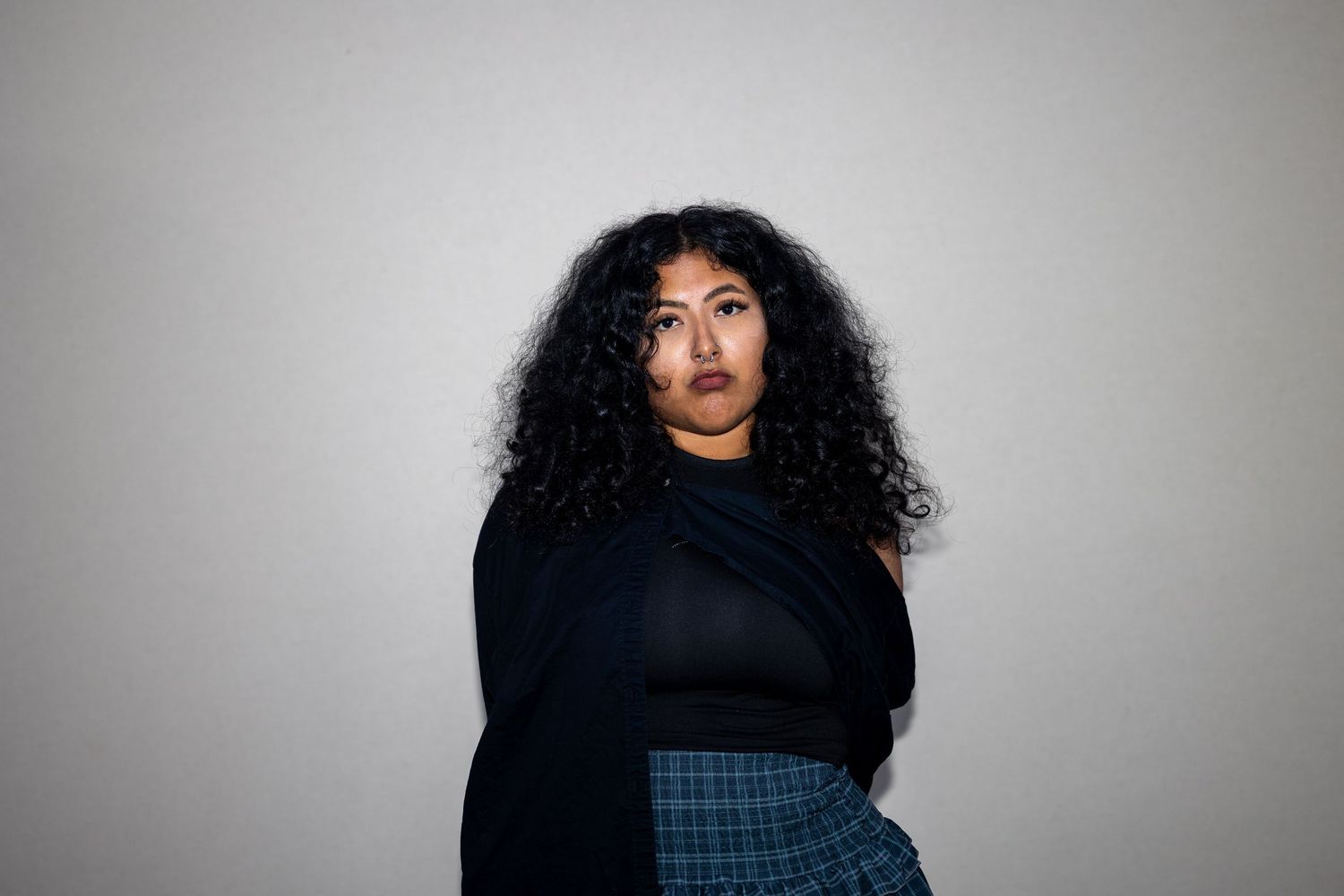 Woman with curly black hair in dark top and plaid skirt poses against gray background.
