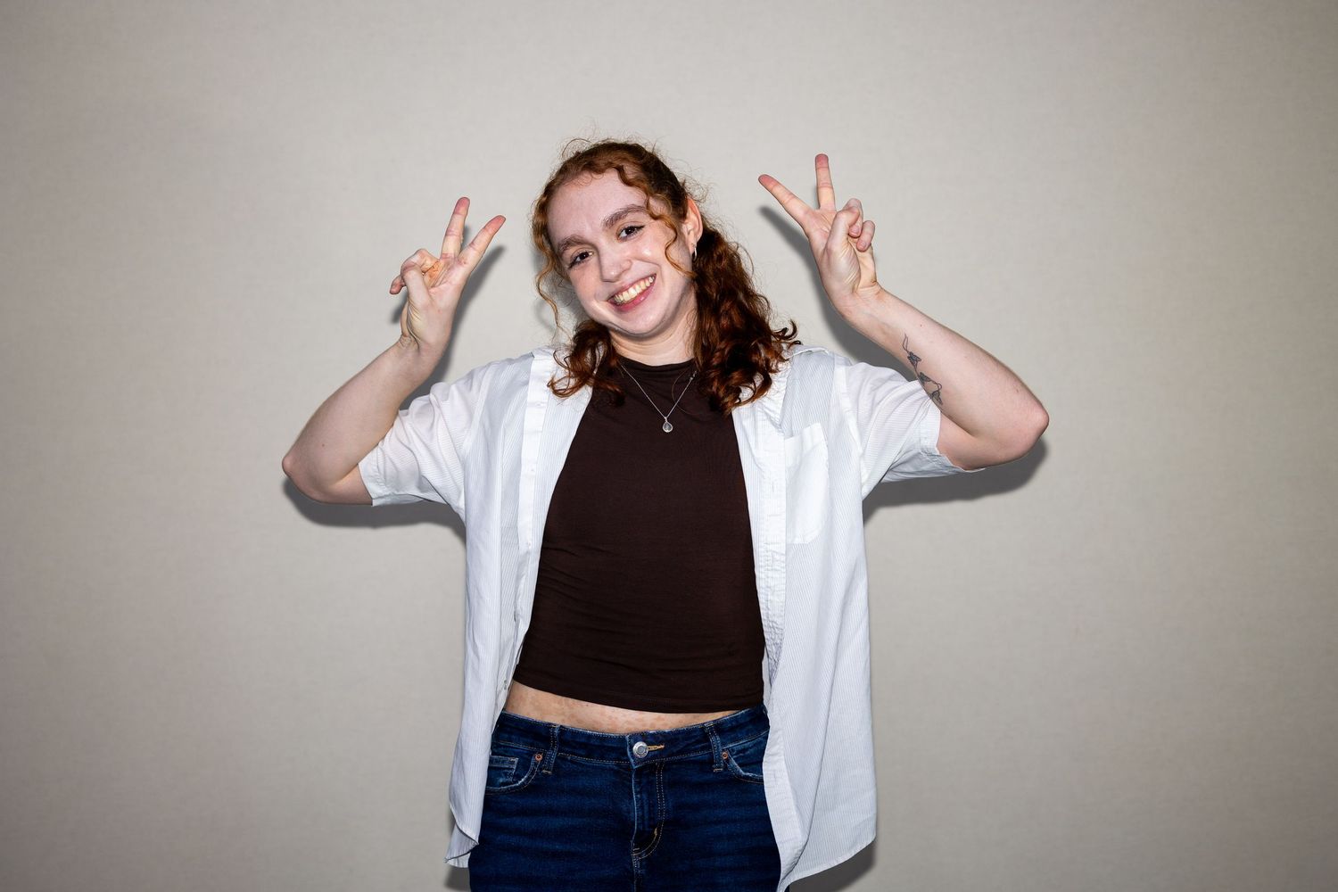 Young woman in white shirt and jeans makes peace signs while smiling against neutral background.