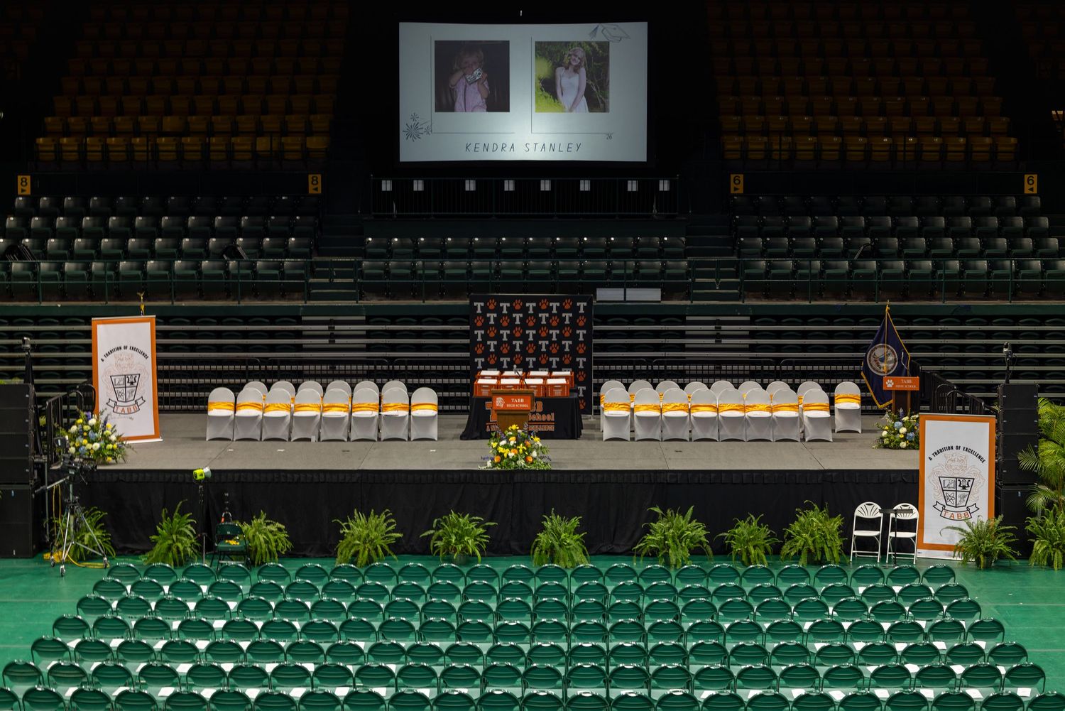 Empty arena memorial service setup with stage, chairs, floral arrangements, and tribute screens displaying photos.