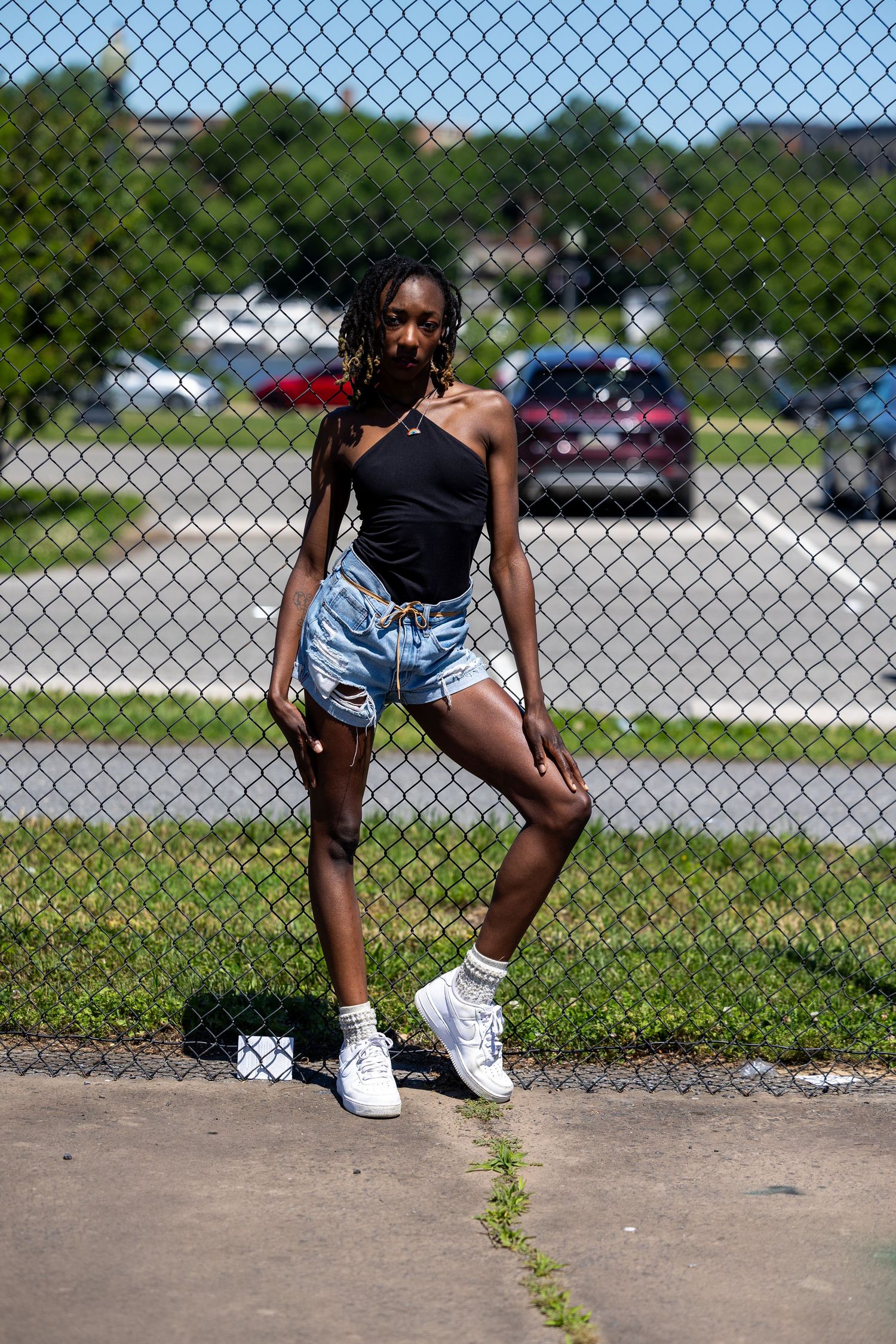Young woman in black top, denim shorts, and white sneakers posing against a chain-link fence outdoors.