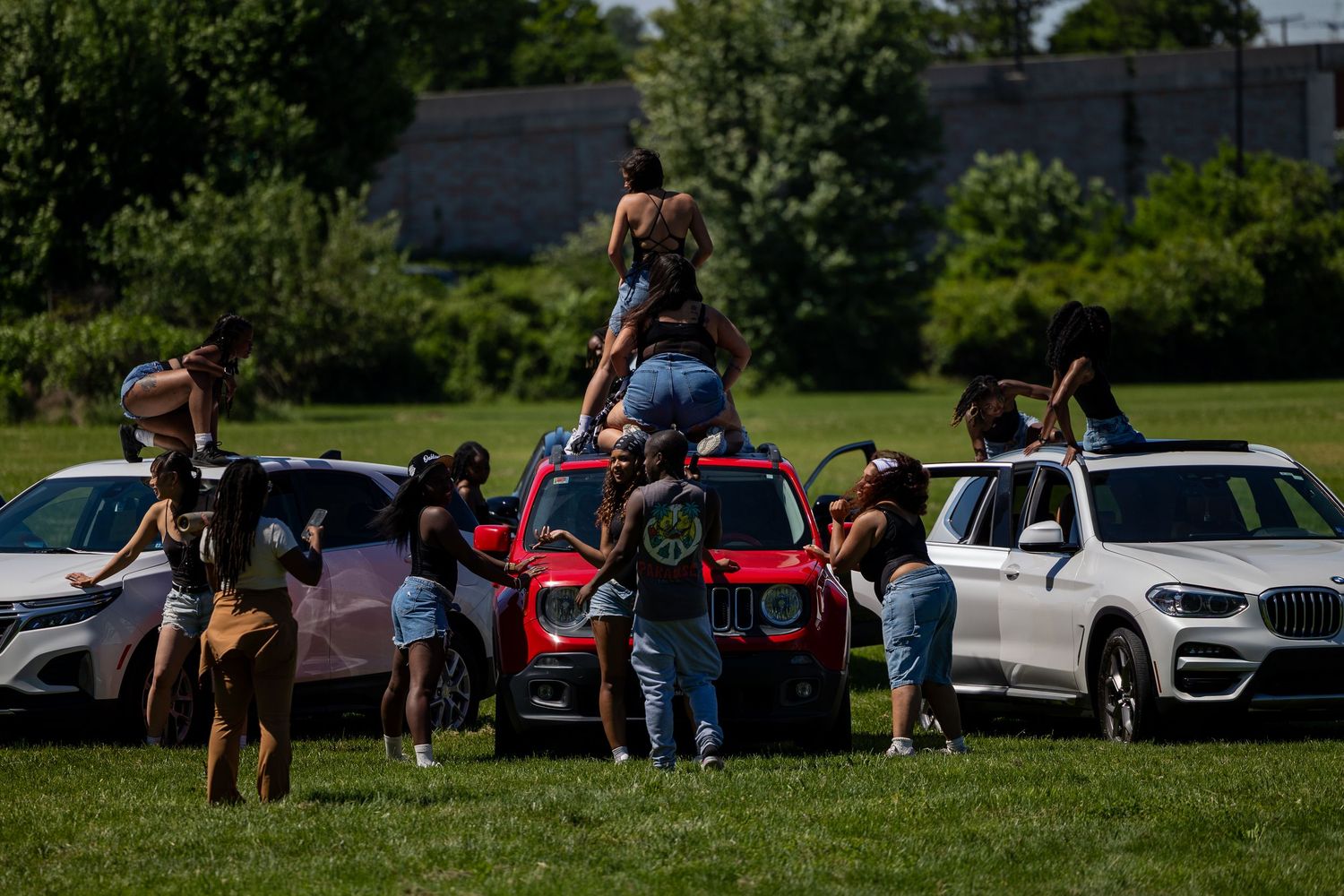 Group of young people gathered around red and white cars on a sunny green field outdoors.
