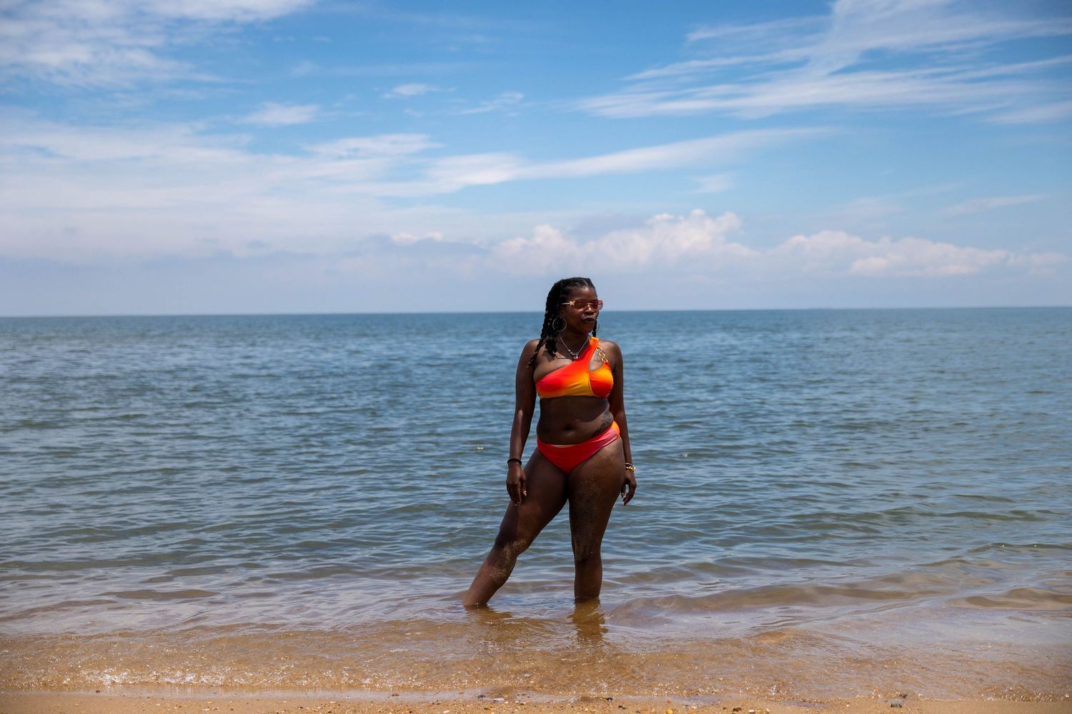 Woman in orange bikini standing in shallow ocean water on a sunny beach with blue sky and clouds.