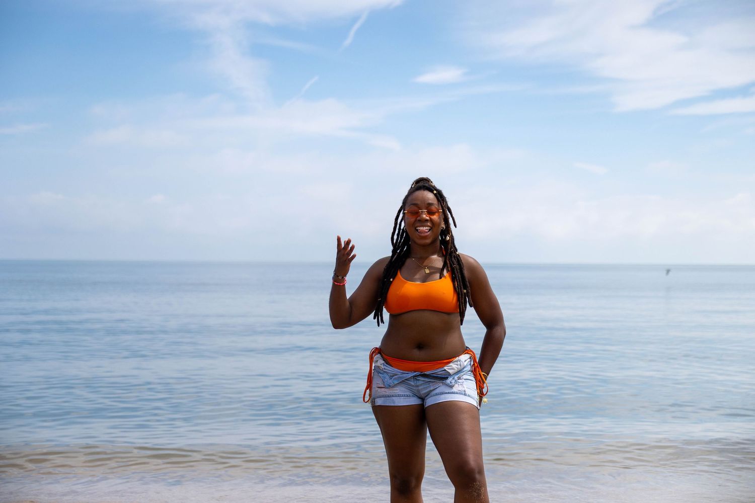 Woman in orange bikini top and denim shorts making peace sign on sunny beach with blue ocean backdrop.