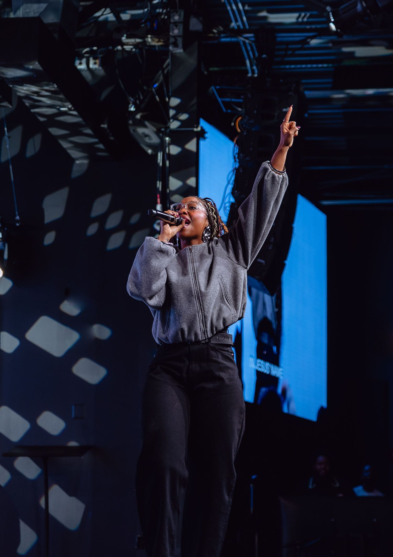 A performer in a grey sweatshirt sings passionately on stage with dramatic lighting and shadows in the background.