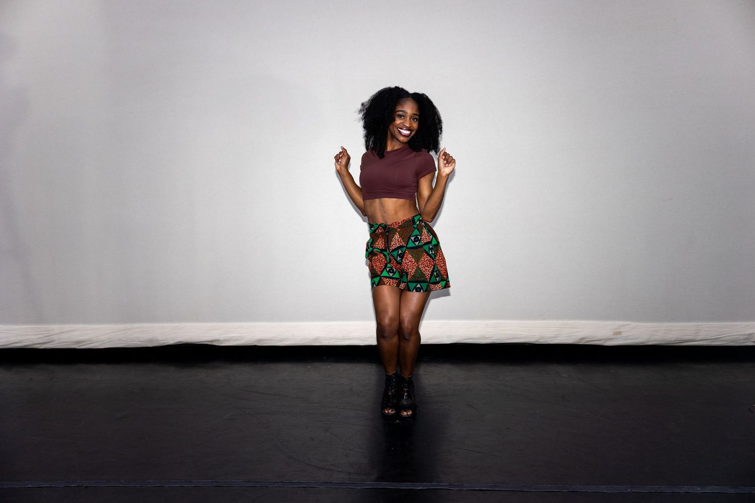 A woman in a vibrant African print skirt and crop top dances joyfully in a studio.