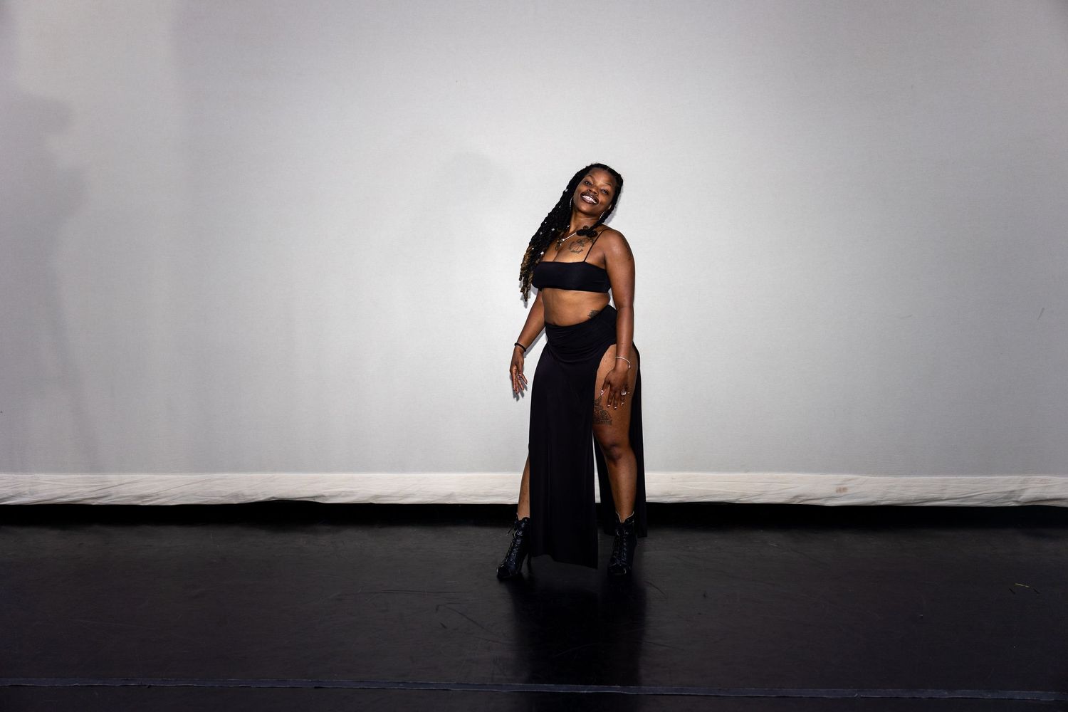 Woman in black crop top and slit maxi skirt poses confidently on dark stage floor against white wall.