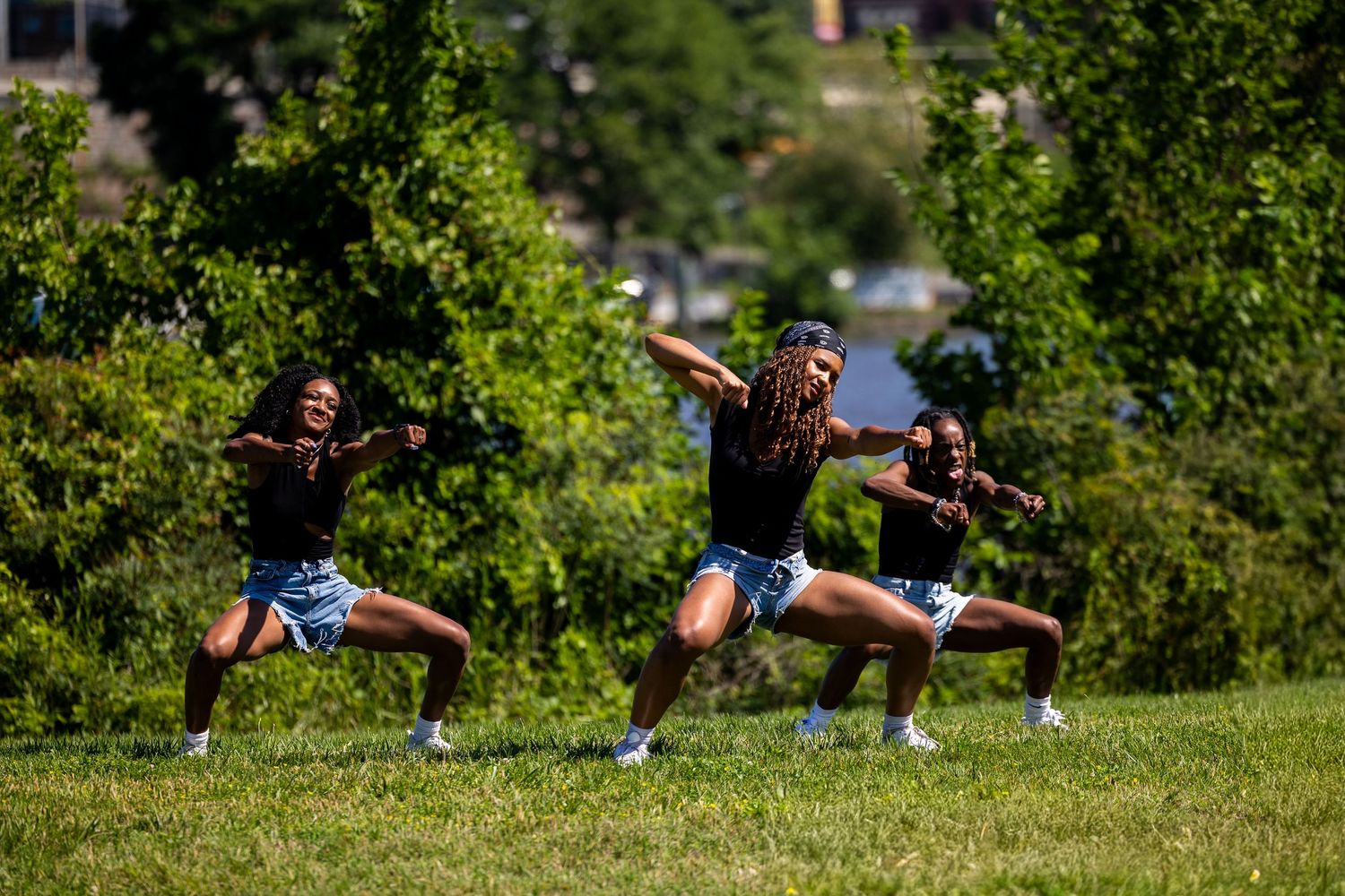 Three women in matching black tops and blue shorts perform synchronized dance moves on a sunny green lawn.