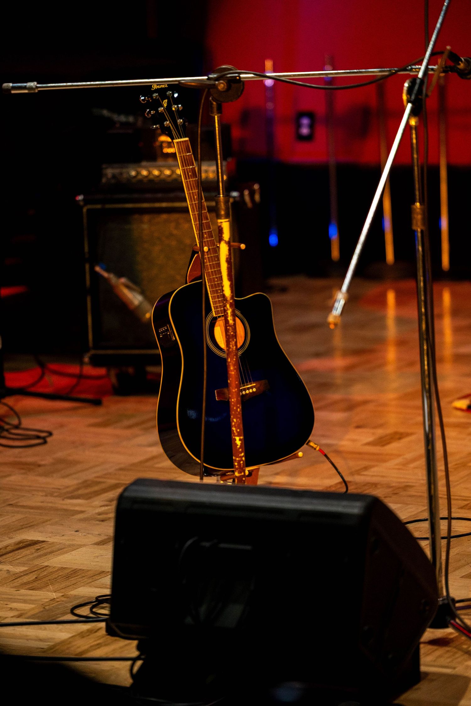 Acoustic guitar on stand with mic stands and amplifiers on a warmly lit wooden stage.