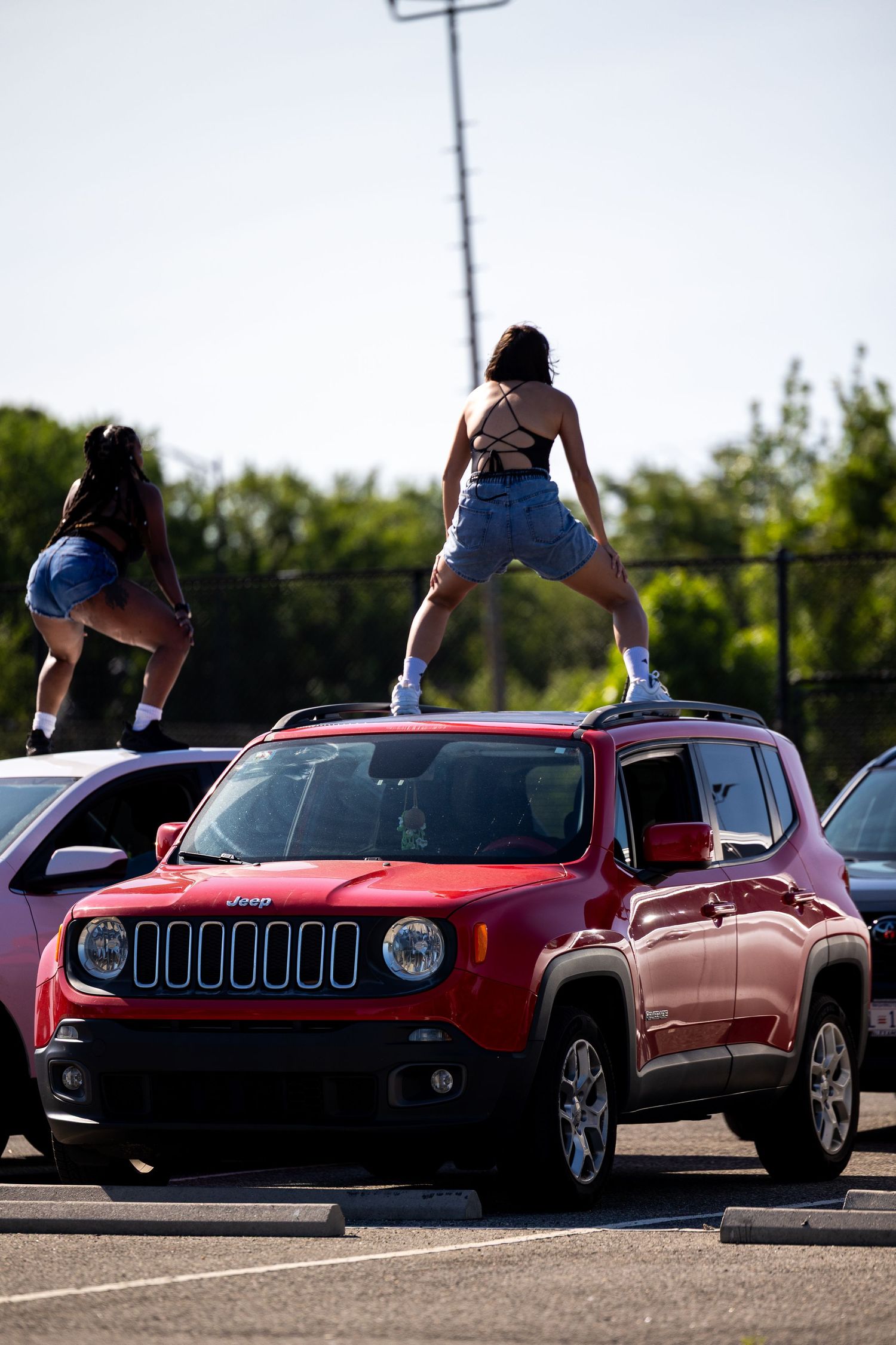 Two women standing on top of a red Jeep Renegade in a sunny parking lot, wearing denim shorts.