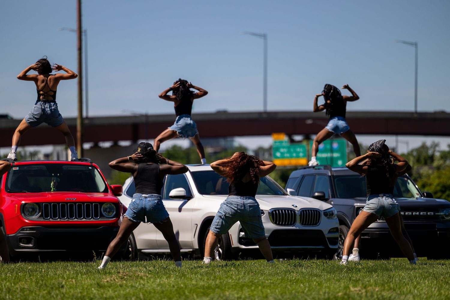 Group of women in denim shorts posing on cars on a highway, striking synchronized poses under a clear blue sky.