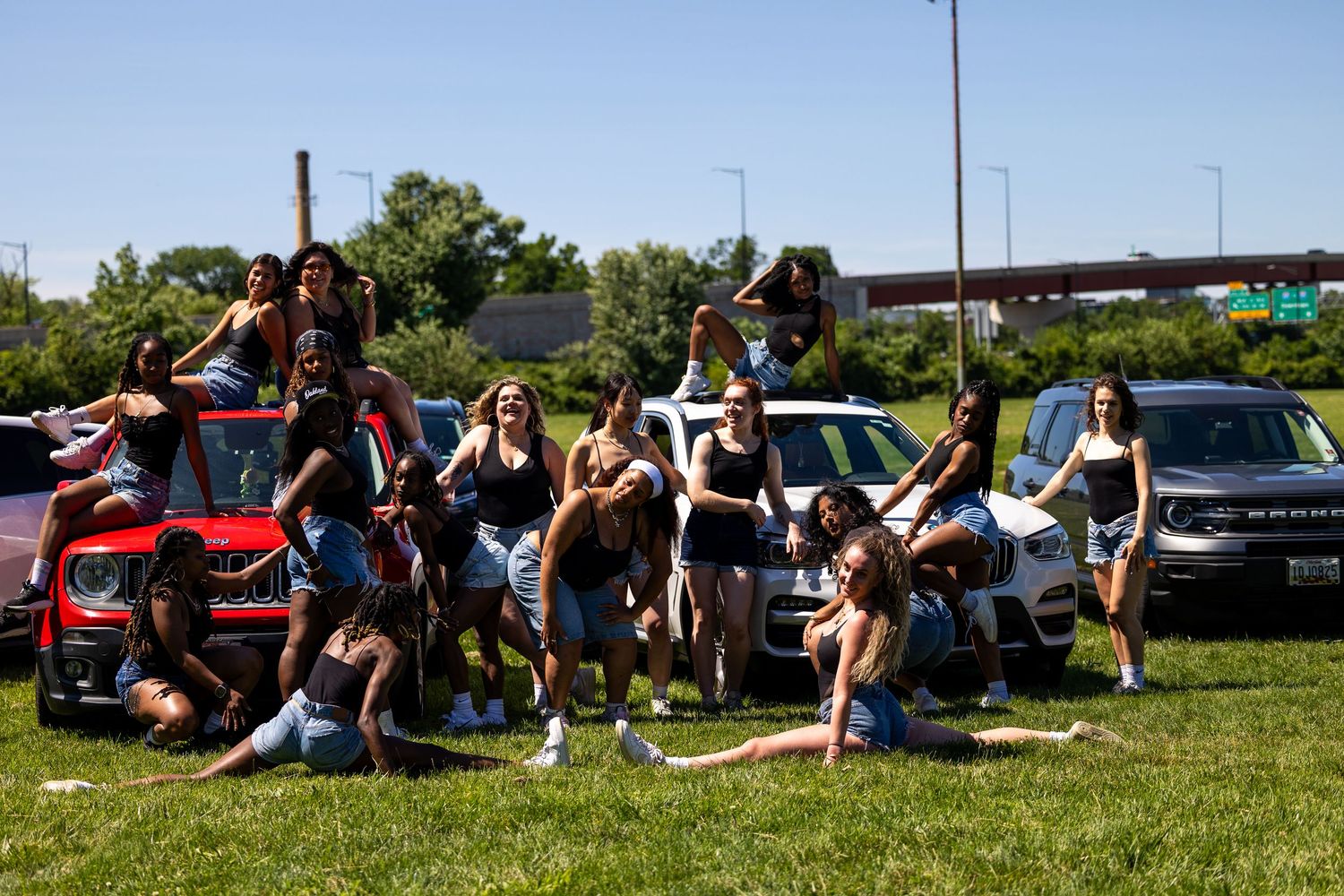 Group of young women posing together on grass near parked cars on a sunny day outdoors.