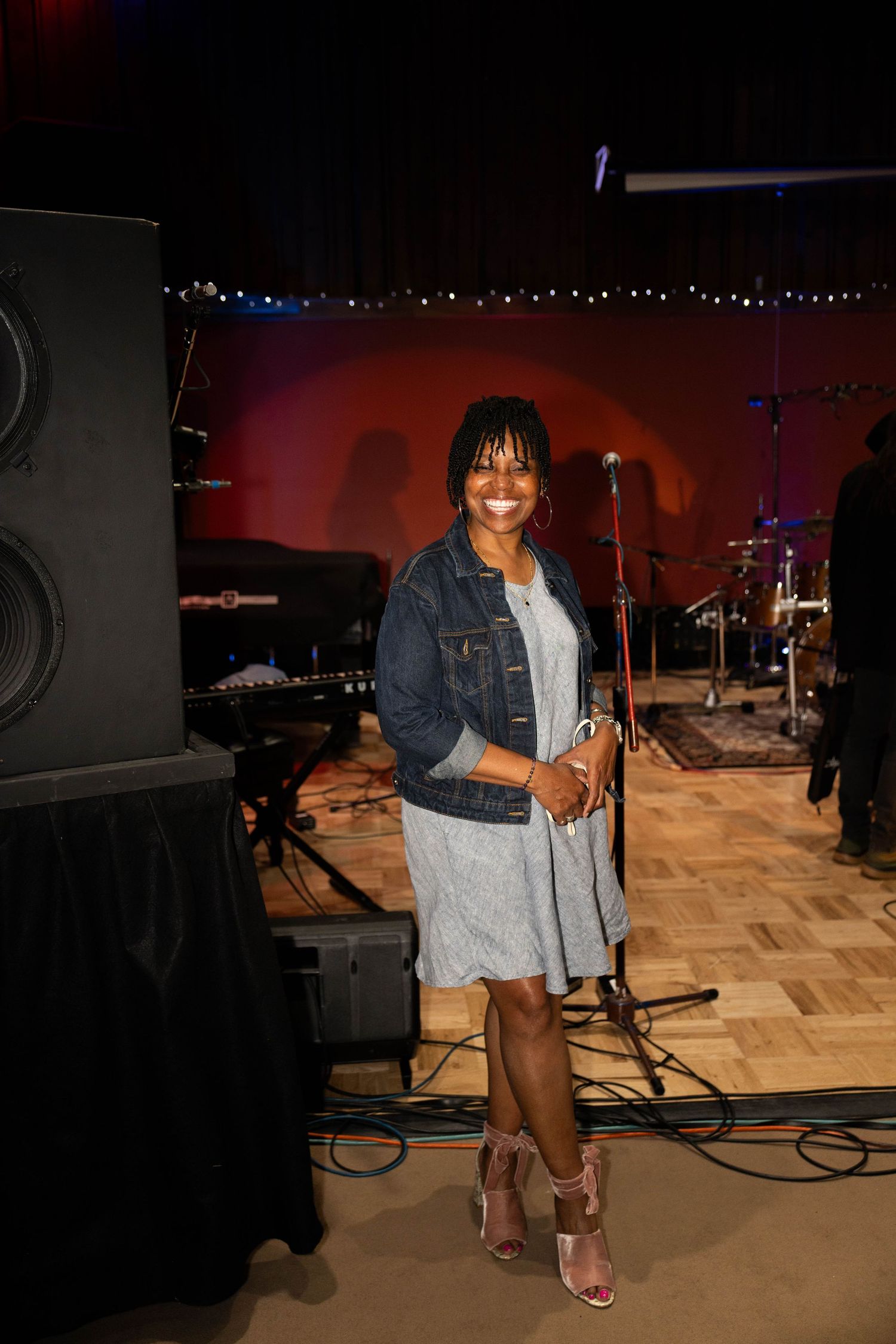 Smiling woman in denim jacket and gray dress stands on stage near speakers and microphones at a music venue.