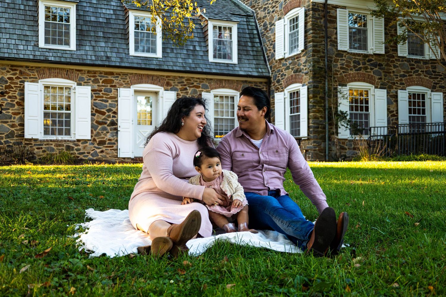 Family of three sitting on blanket in grass before stone house on sunny fall day.