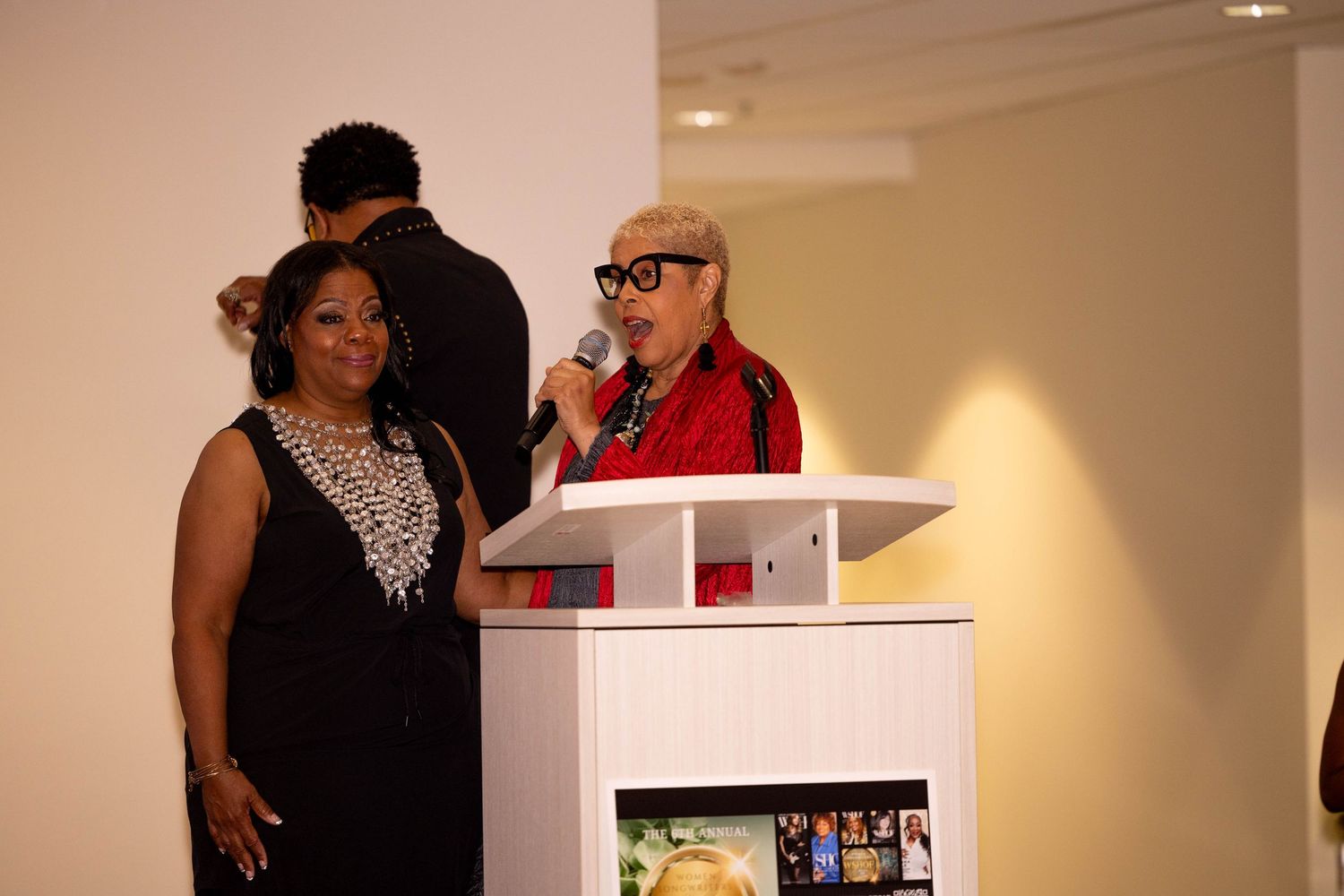Woman in red coat speaks at white podium beside woman in black dress at formal event.