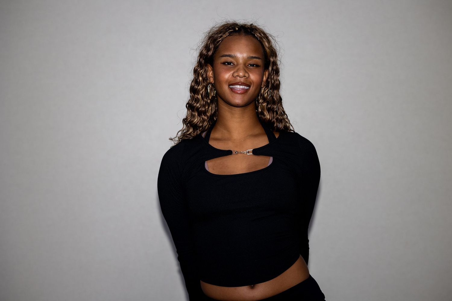 A young woman with highlighted curly hair wearing a black cutout crop top smiles confidently.