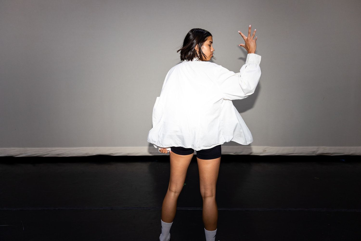 Woman in white oversized jacket and black shorts poses against gray wall, facing away from camera.
